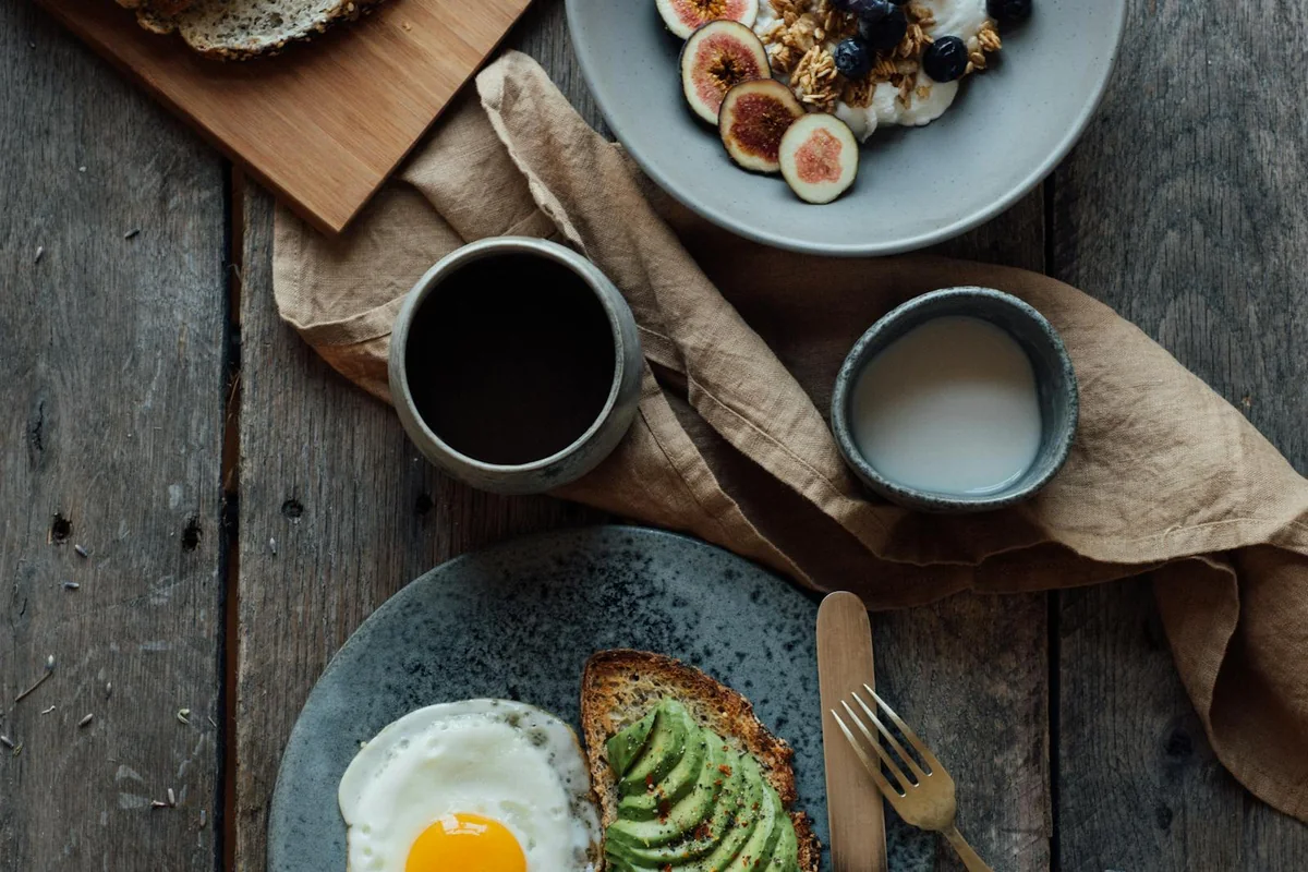 Desayuno con huevo, tostada de aguacate, fruta y café en mesa de madera